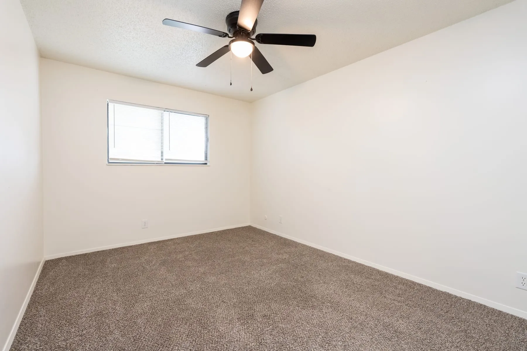 Second sleeping area in a two bedroom home, carpeted with a window and ceiling fan.